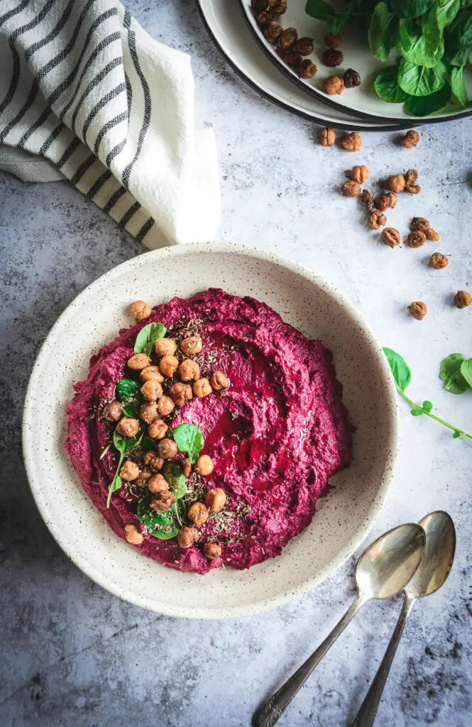 Beet hummus in bowl topped with greens and crispy chickpeas, with white and black napkin and two spoons