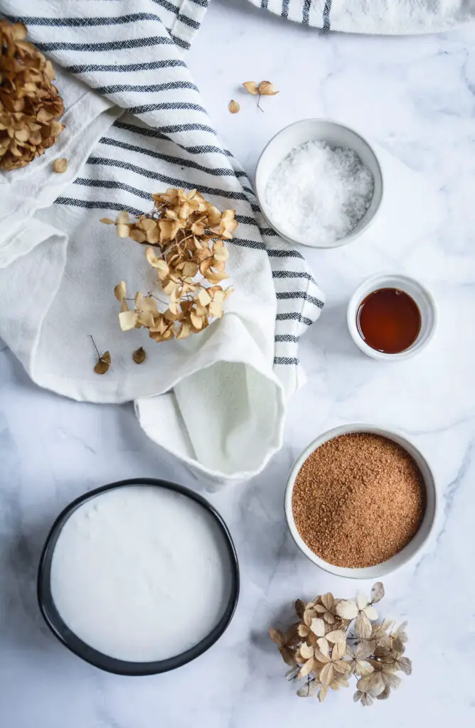 Coconut cream in bowl, coconut sugar in bowl, vanilla extract in bowl, salt in bowl, napkin and dried flowers 