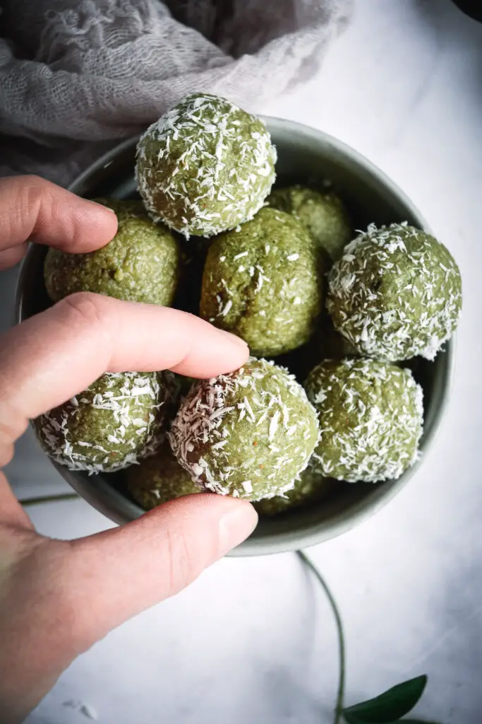 Coconut Cashew Matcha Honey Energy Bites in bowl being picked up by thumb and index finger