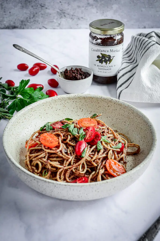 Moroccan Tapenade Spaghetti with Tomatoes and Herbs in bowl with jar of tapenade, bowl of tapenade, herbs and tomatoes