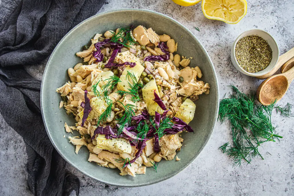 White bean tuna salad with artichoke hearts in bowl with dark colored napkin and lemons 