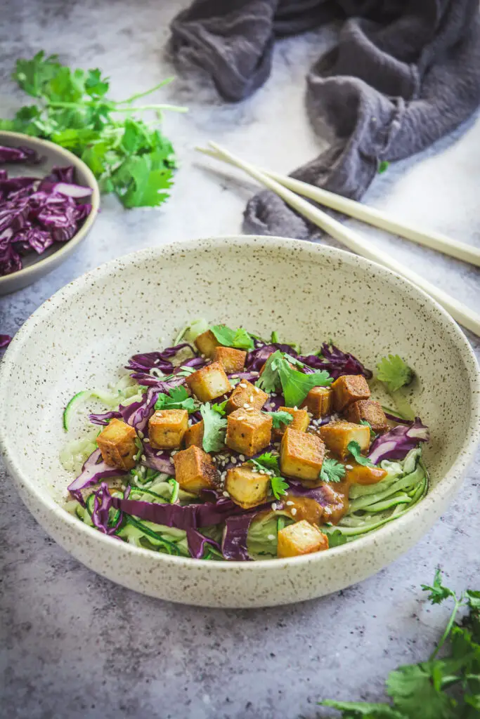 Zoodles with Thai Peanut Sauce and Tofu on table with cilantro and chopsticks, red cabbage and napkin