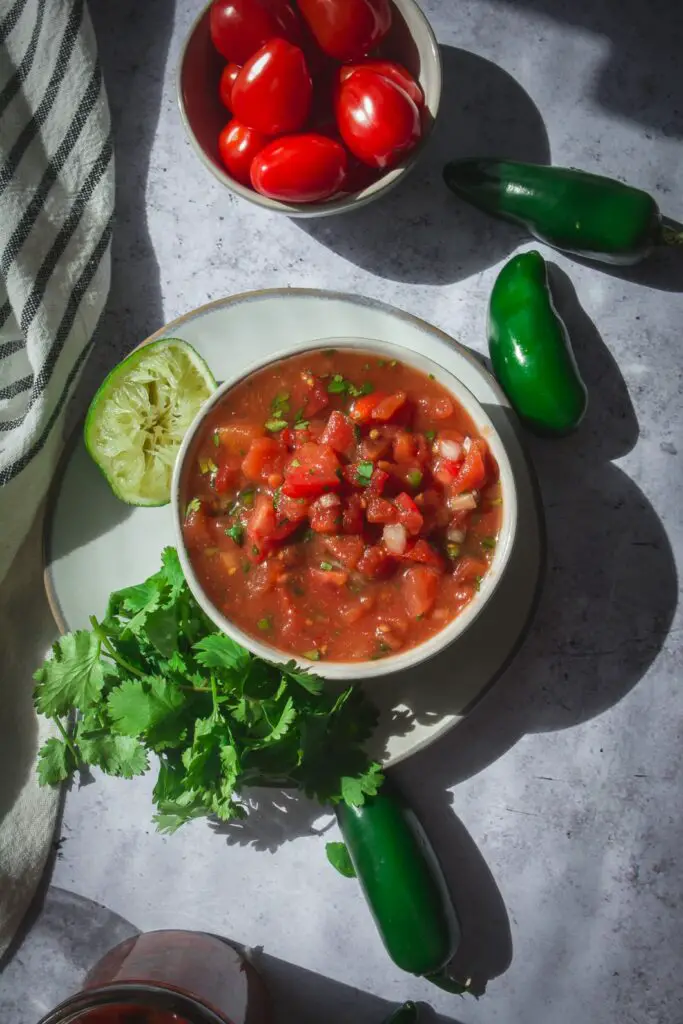 homemade salsa in bowl on plate with tomatoes, jalapenos, cilantro and lime next to bowl.