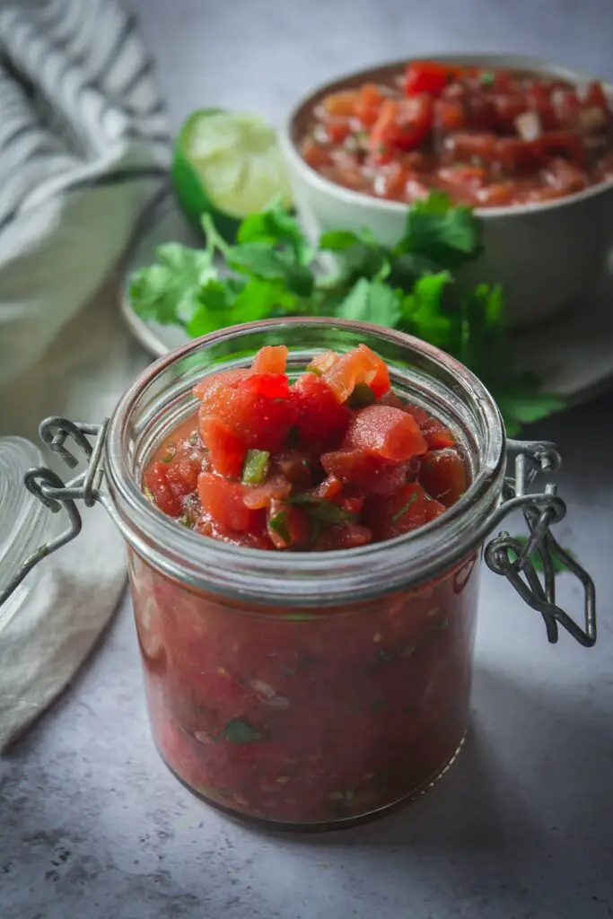 Homemade salsa in jar with cilantro and bowl of salsa in the background