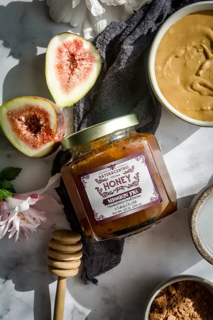 jar of honey, honey scooper, peanut butter bowl, halves of fresh figs and hibiscus flower