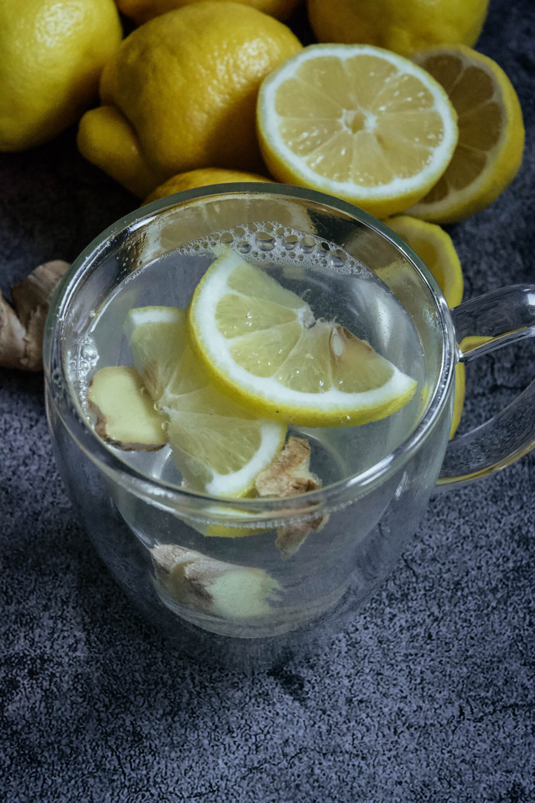 Lemon water in clear mug with lemon slices and ginger and lemons in the background