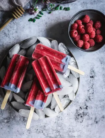 4 raspberry popsicles on a plate of ice cubes and a bowl of raspberries next to it