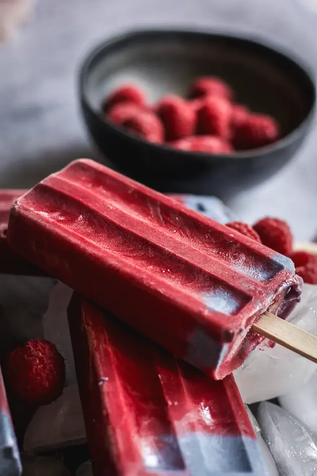 raspberry popsicles on a plate of ice cubes and a bowl of raspberries next to it