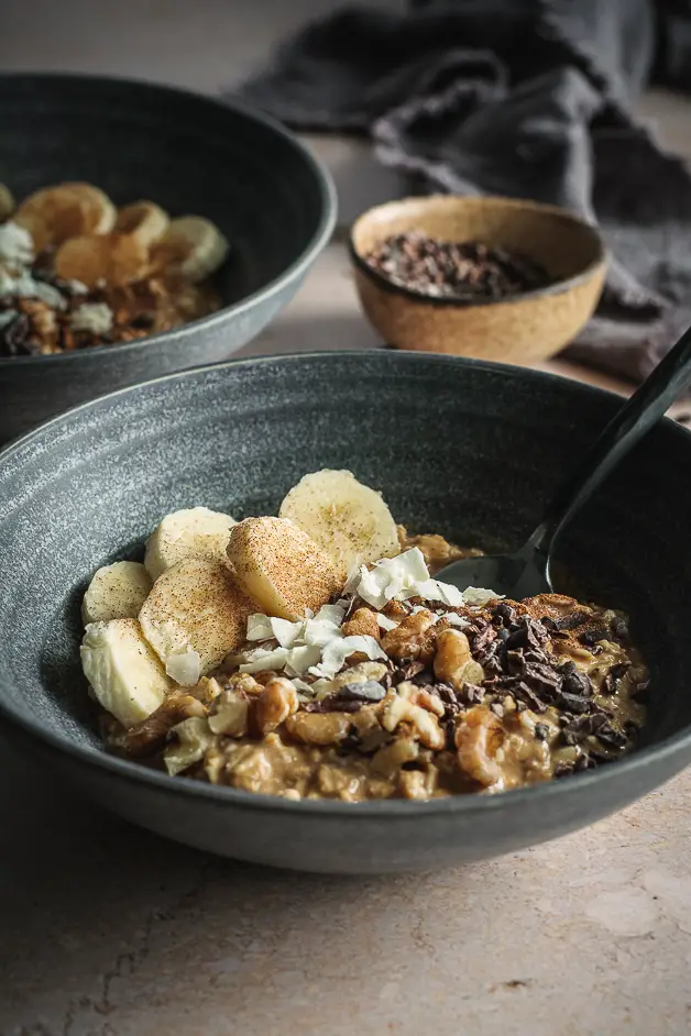pumpkin oatmeal in blue bowl with bananas, walnuts, coconut flakes and cacao nibs and a spoon