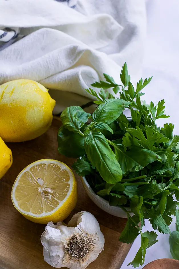 fresh herbs on a cutting board with a zested lemon, a lemon half and a head of garlic