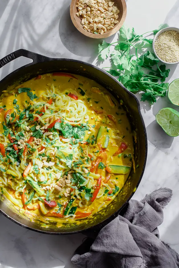 a skillet of thai peanut sauce with noodles, lime halves, peanuts and sesame seeds