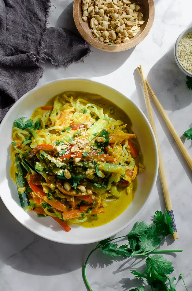 bowl of noodles with peanut sauce on table with chopsticks, a grey napkin, cut up lime, a bowl of peanuts and a bowl of sesame seeds