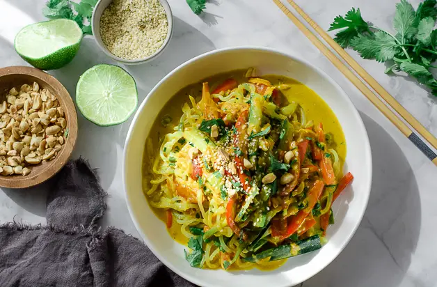 bowl of noodles with peanut sauce on table with chopsticks, a grey napkin, cut up lime, a bowl of peanuts and a bowl of sesame seeds