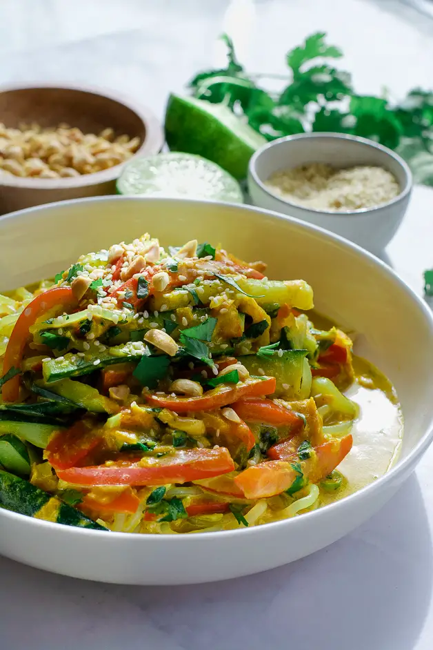 bowl of noodles with peanut sauce on table with chopsticks, a grey napkin, cut up lime, a bowl of peanuts and a bowl of sesame seeds