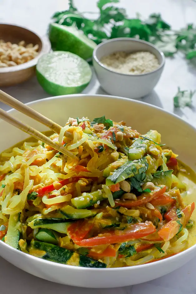 bowl of noodles with peanut sauce on table with chopsticks, cut up lime, a bowl of peanuts and a bowl of sesame seeds