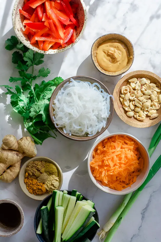 a small bowl of grated carrot, a bowl of sliced red peppers, sliced zucchini, a bowl of spices, a small bowl of peanut butter, a bowl of white noodles, cilantro bunch and a ginger root