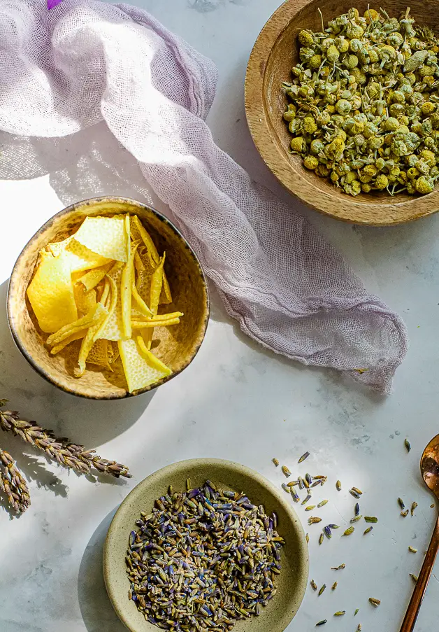a bowl or lemon rind, a bowl of lavender flowers, a bowl of chamomile flowers on a table