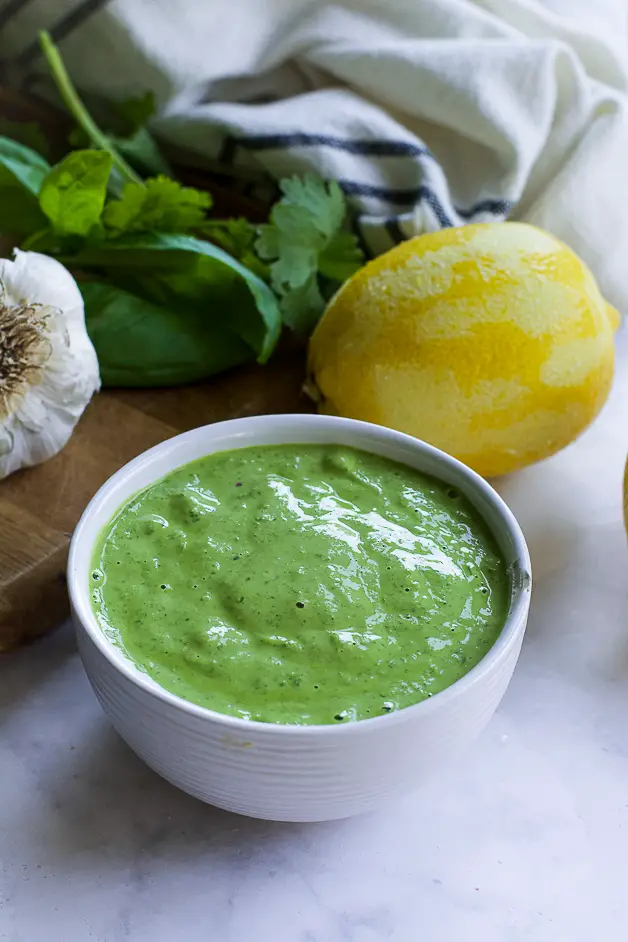 a bowl with green sauce with a small wooden spoon on a table. next to it a lemon, herbs and a head of garlic on a cutting board. 