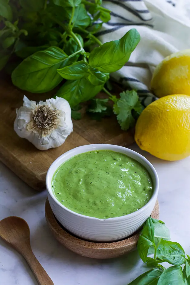 a bowl with green sauce with a small wooden spoon on a table. next to it a lemon, herbs and a head of garlic on a cutting board. 