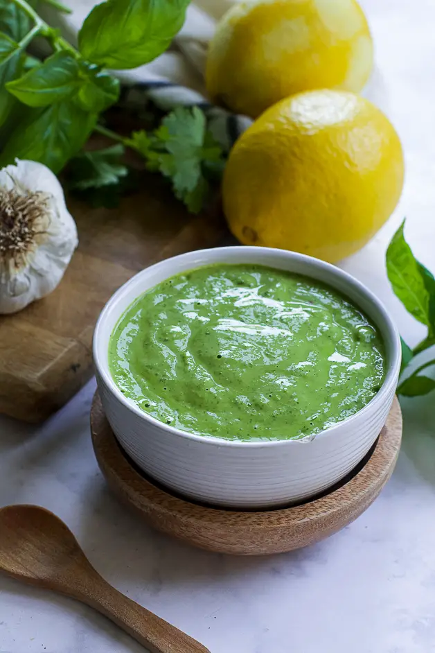 a bowl with green sauce with a small wooden spoon on a table. next to it a lemon, herbs and a head of garlic on a cutting board. 