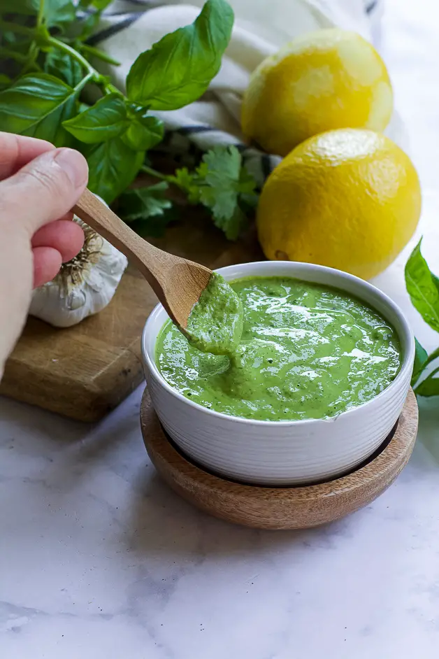 a bowl with green sauce with a small wooden spoon on a table. next to it a lemon, herbs and a head of garlic on a cutting board. 