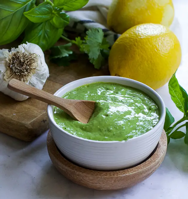 a bowl with green sauce with a small wooden spoon on a table. next to it a lemon, herbs and a head of garlic on a cutting board. 