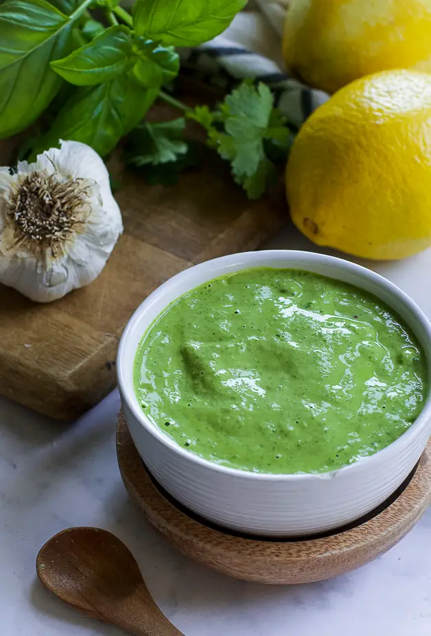 a bowl with green sauce with a small wooden spoon on a table. next to it a lemon, herbs and a head of garlic on a cutting board. 