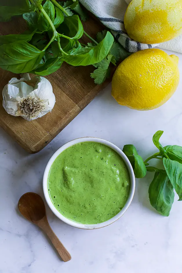 a bowl with green sauce with a small wooden spoon on a table. next to it a lemon, herbs and a head of garlic on a cutting board. 