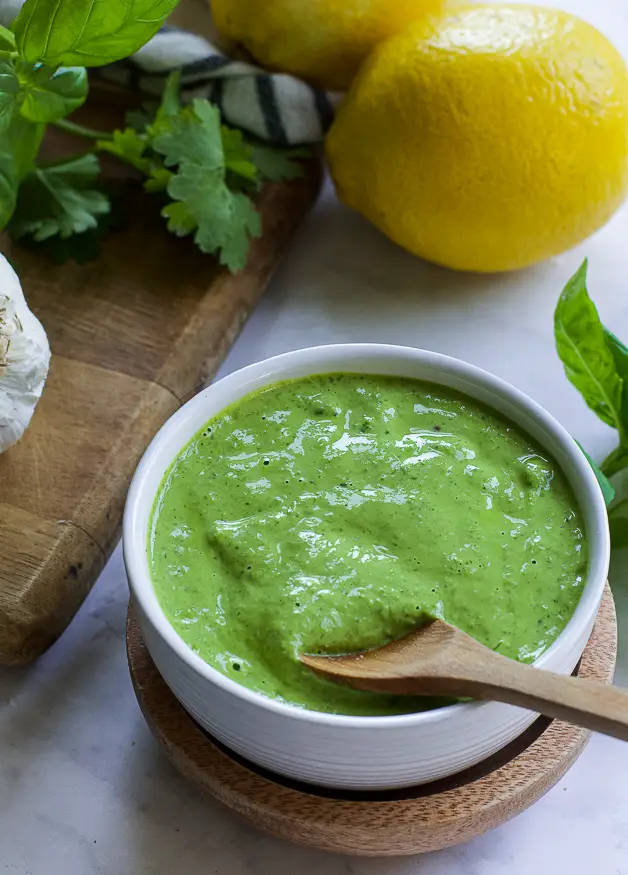 a bowl with green sauce with a small wooden spoon on a table. next to it a lemon, herbs and a head of garlic on a cutting board. 