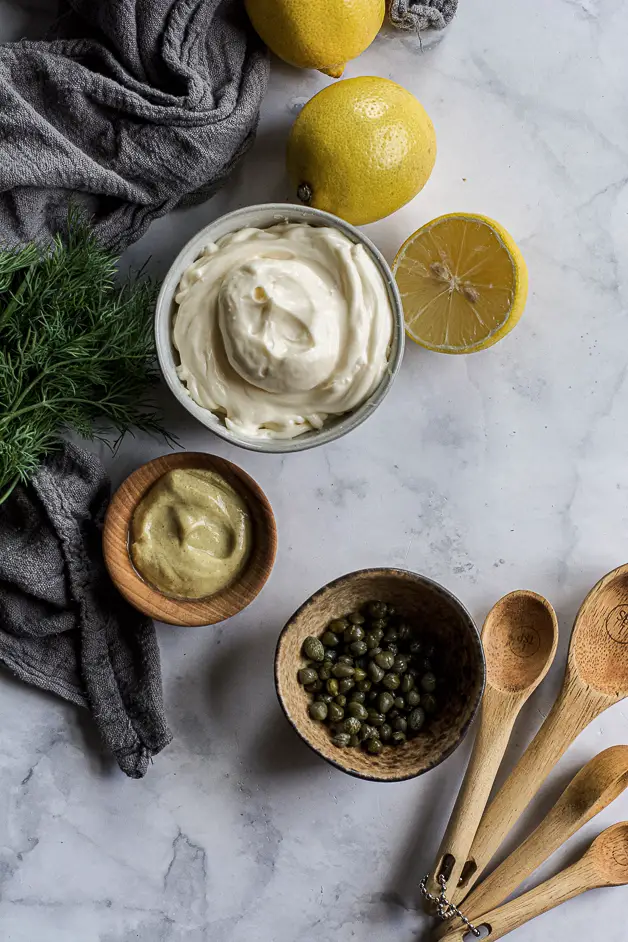 white creamy substance in a bowl, yellowish creamy mustard in a bowl, small capers in a bowl, herbs and lemon halves with wooden spoons on a table