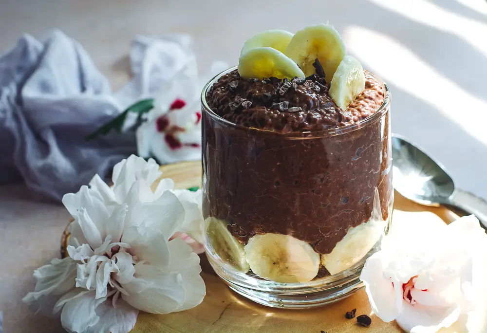 Chocolate chia pudding in a glass topped with bananas and flowers next to it