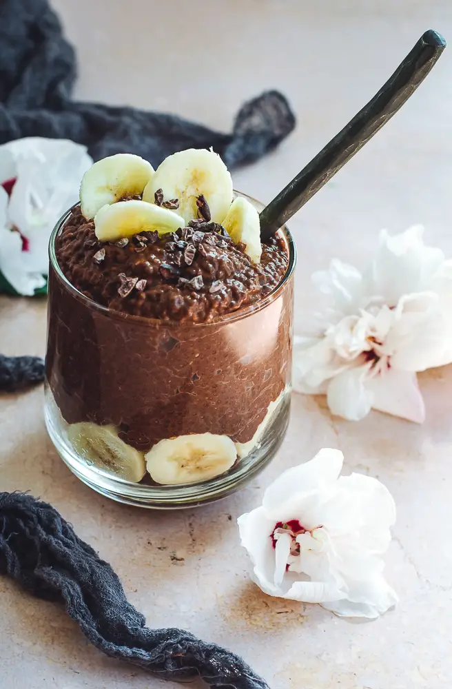 Chocolate chia pudding in a glass topped with bananas and flowers next to it
