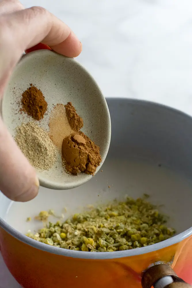 spices being added to a pan with white liquid and chamomile flowers