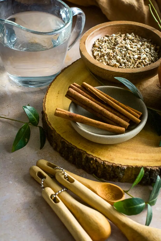 cinnamon sticks on a small plate, chopped roots in a small bowl, clean liquid in a glass