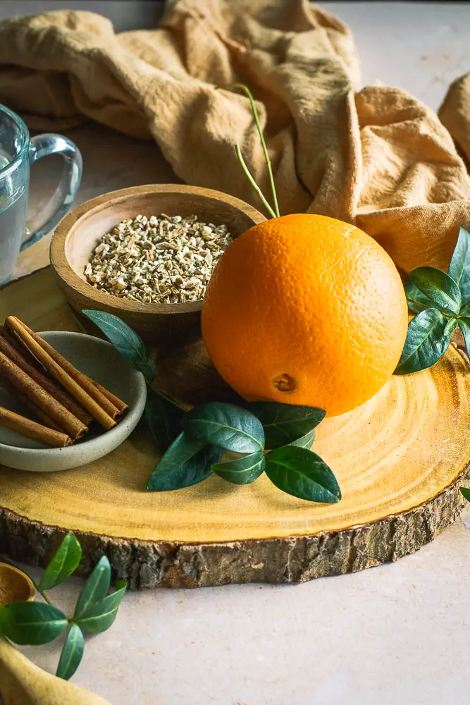 cinnamon sticks on a small plate, chopped roots in a small bowl, clean liquid in a glass and an orange