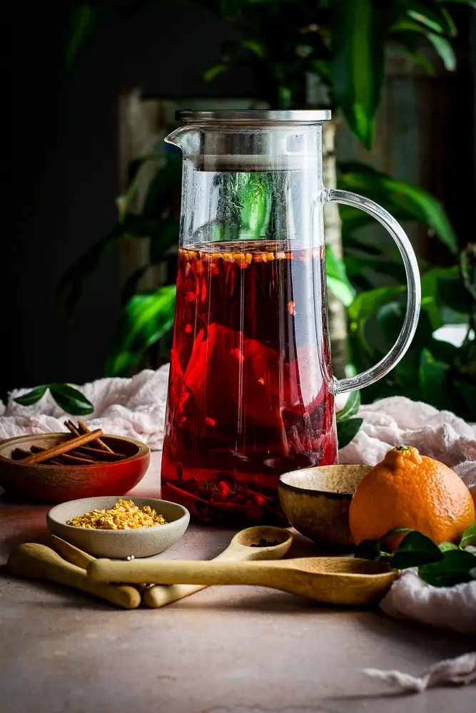 tall tea pitcher with red tea, small bowl of dried orange, dried hibiscus and cinnamon sticks