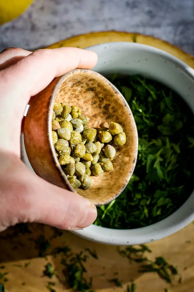 capers being added to a bowl of herbs