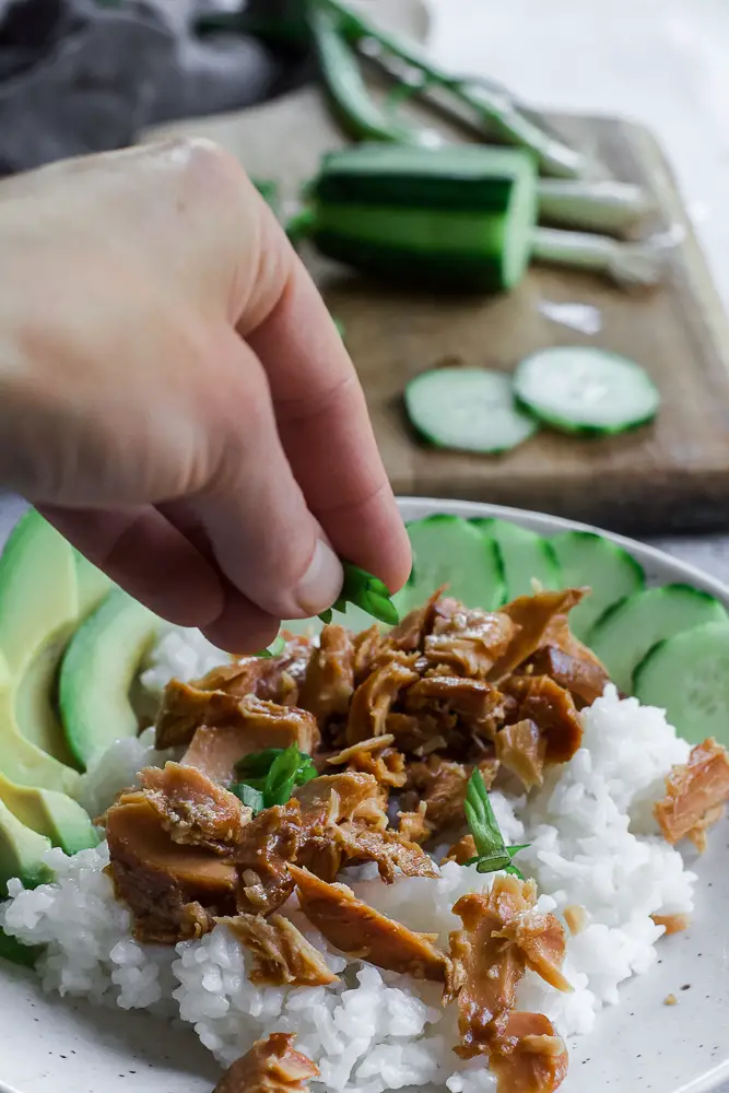 Bowl of rice and tunafish in a white bowl with sliced green cucumbers and sliced green avocado.