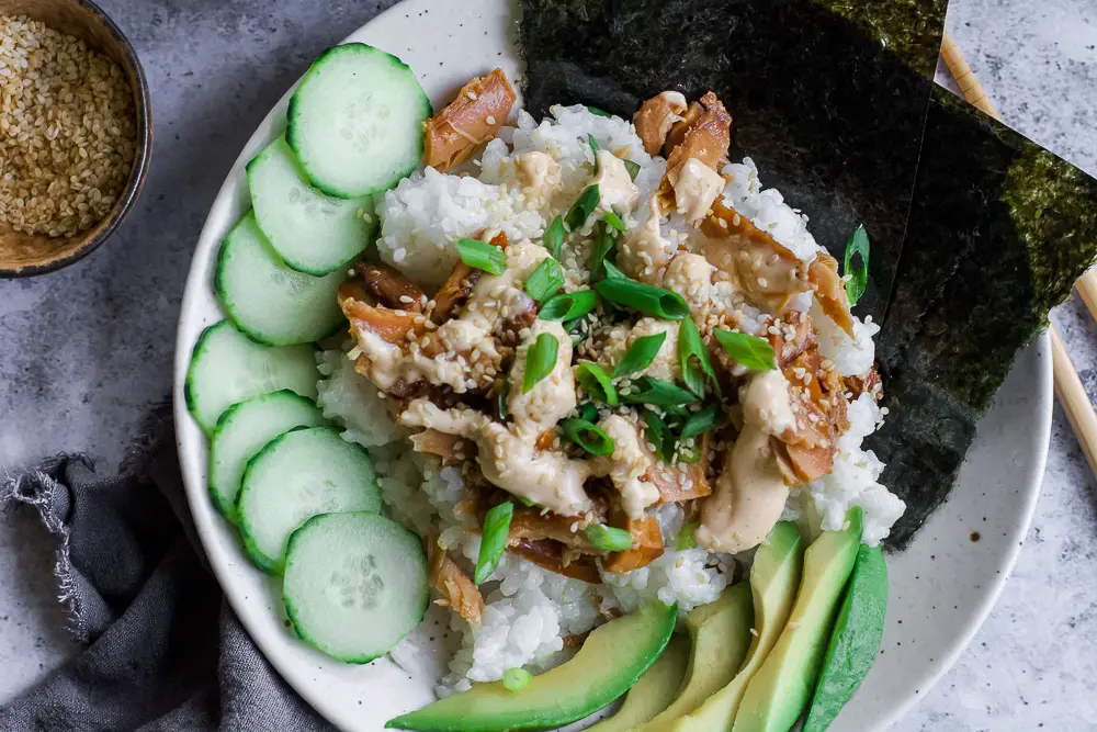 Bowl of rice and tunafish in a white bowl with sliced green cucumbers and sliced green avocado.