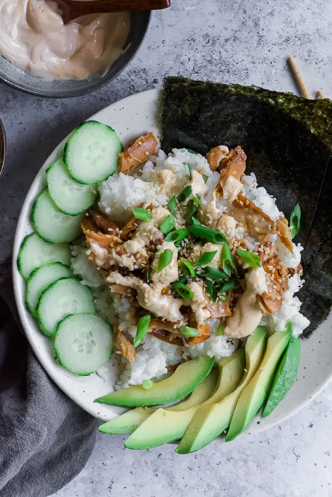 Bowl of rice and tunafish in a white bowl with sliced green cucumbers and sliced green avocado.