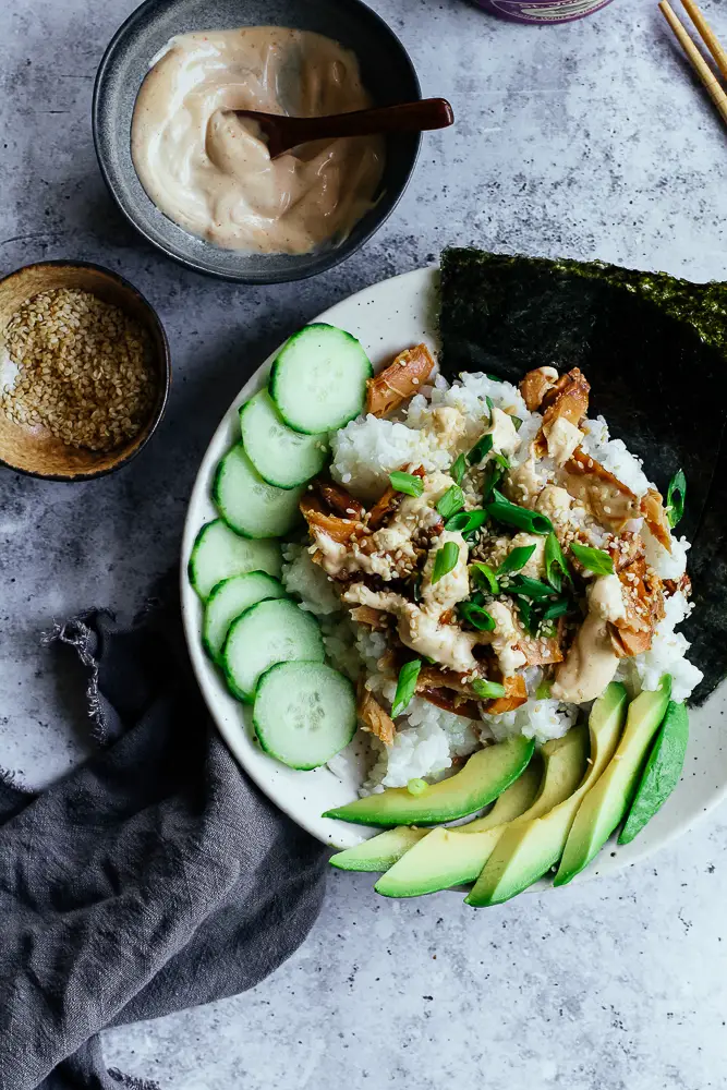 Bowl of rice and tunafish in a white bowl with sliced green cucumbers and sliced green avocado.
