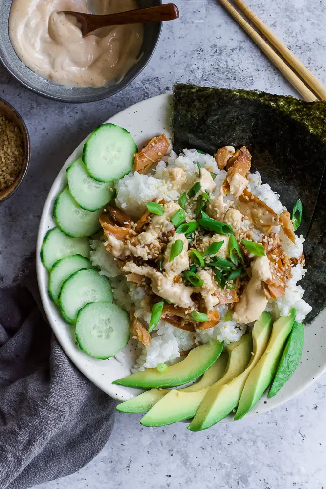Bowl of rice and tunafish in a white bowl with sliced green cucumbers and sliced green avocado.
