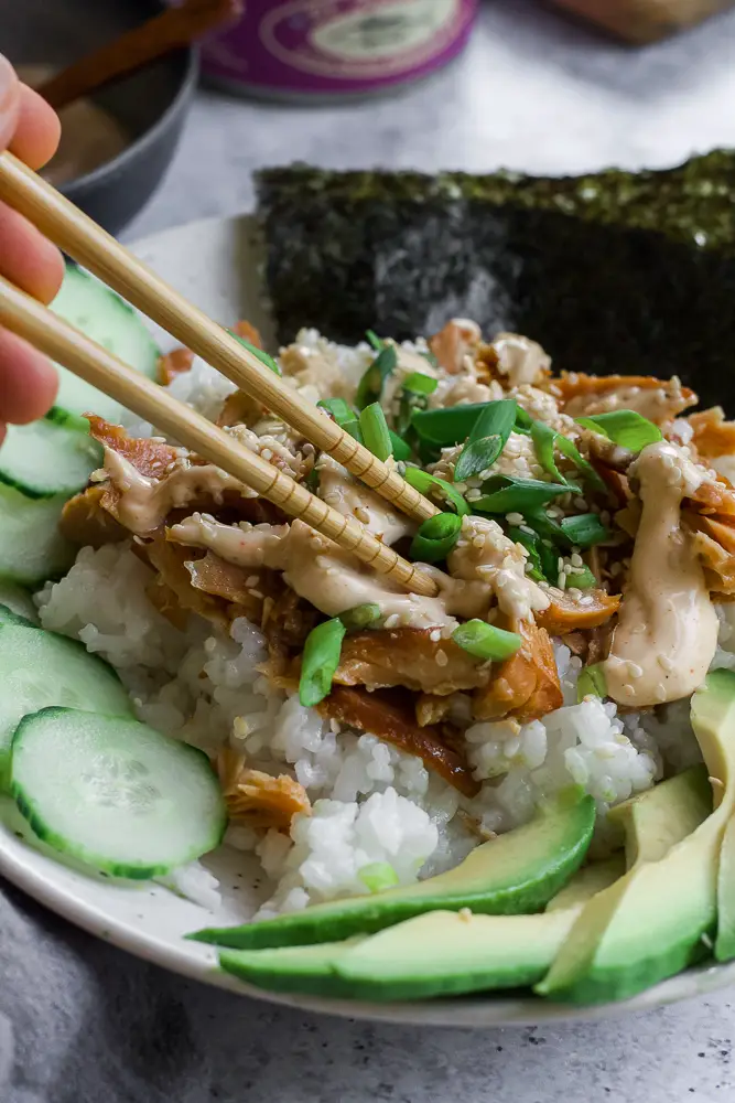 Bowl of rice and tunafish in a white bowl with sliced green cucumbers and sliced green avocado.